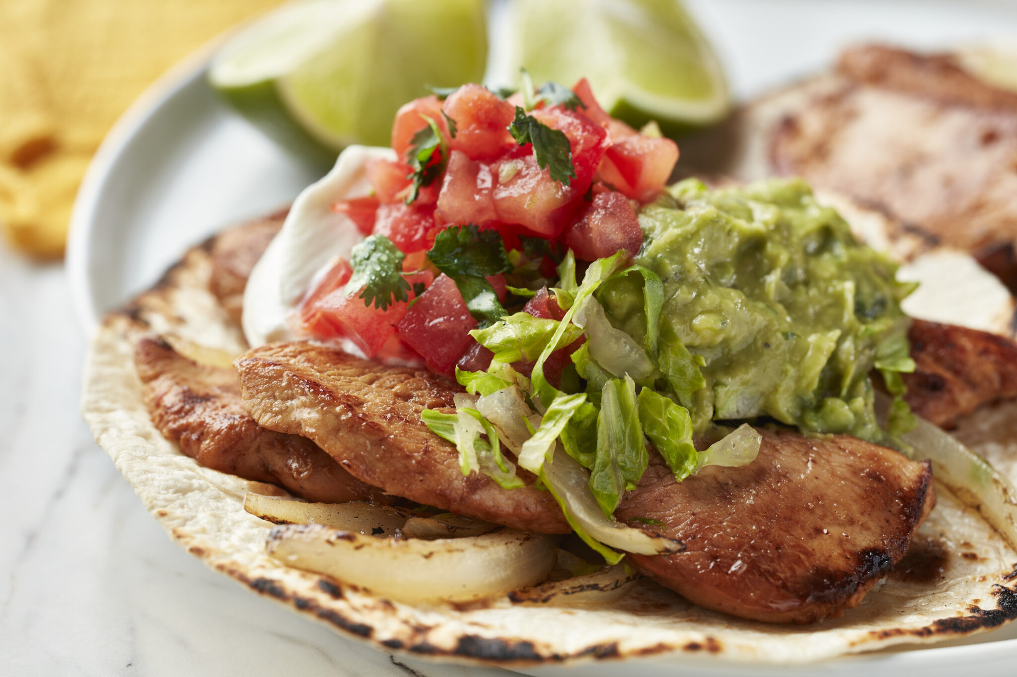 Chicken fajitas on a warm, slightly charred tortilla, topped with fresh guacamole, diced tomatoes, cilantro, shredded lettuce, and a dollop of sour cream, served with lime wedges for garnish.