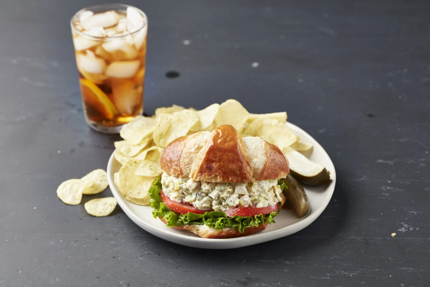 A flaky croissant sandwich filled with creamy chicken salad, lettuce, and tomatoes, served with potato chips and iced tea.