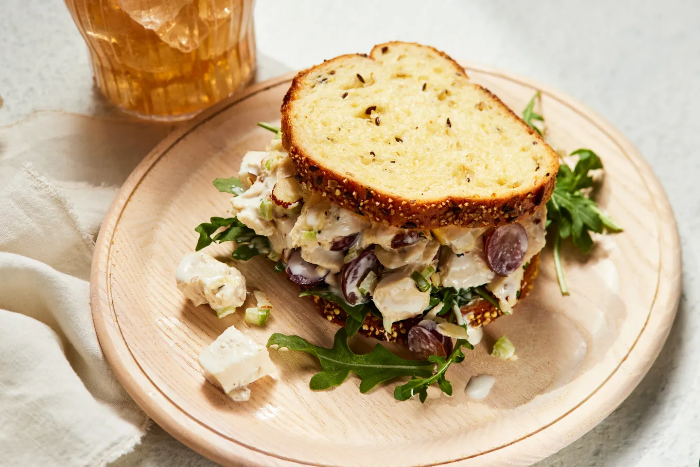 A gourmet chicken salad sandwich with grapes and greens, served on seeded bread with a side of arugula and scattered ingredients on a wooden plate.