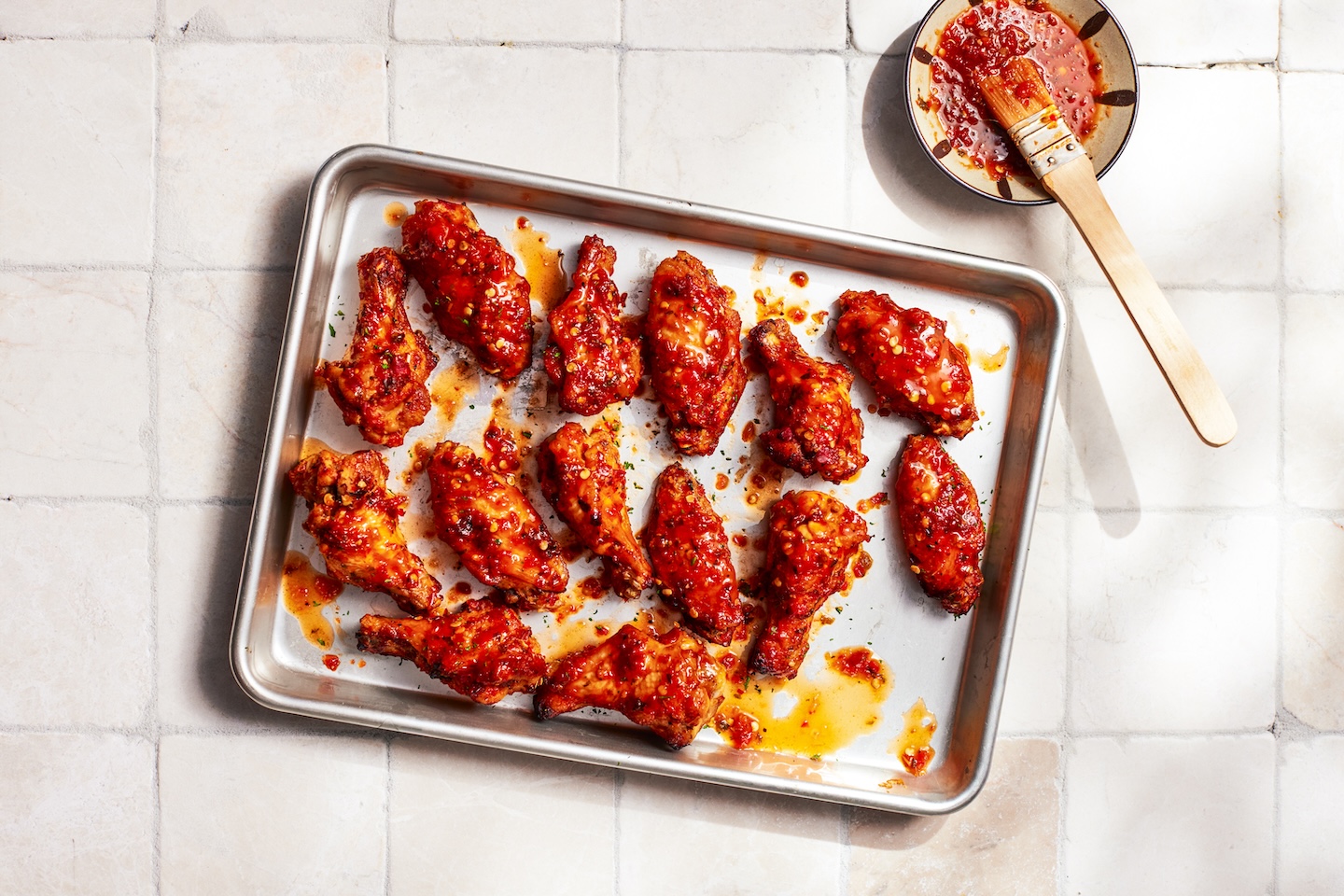 A baking sheet filled with sweet, tangy chicken wings coated in a red pepper sauce with visible chili flakes.
